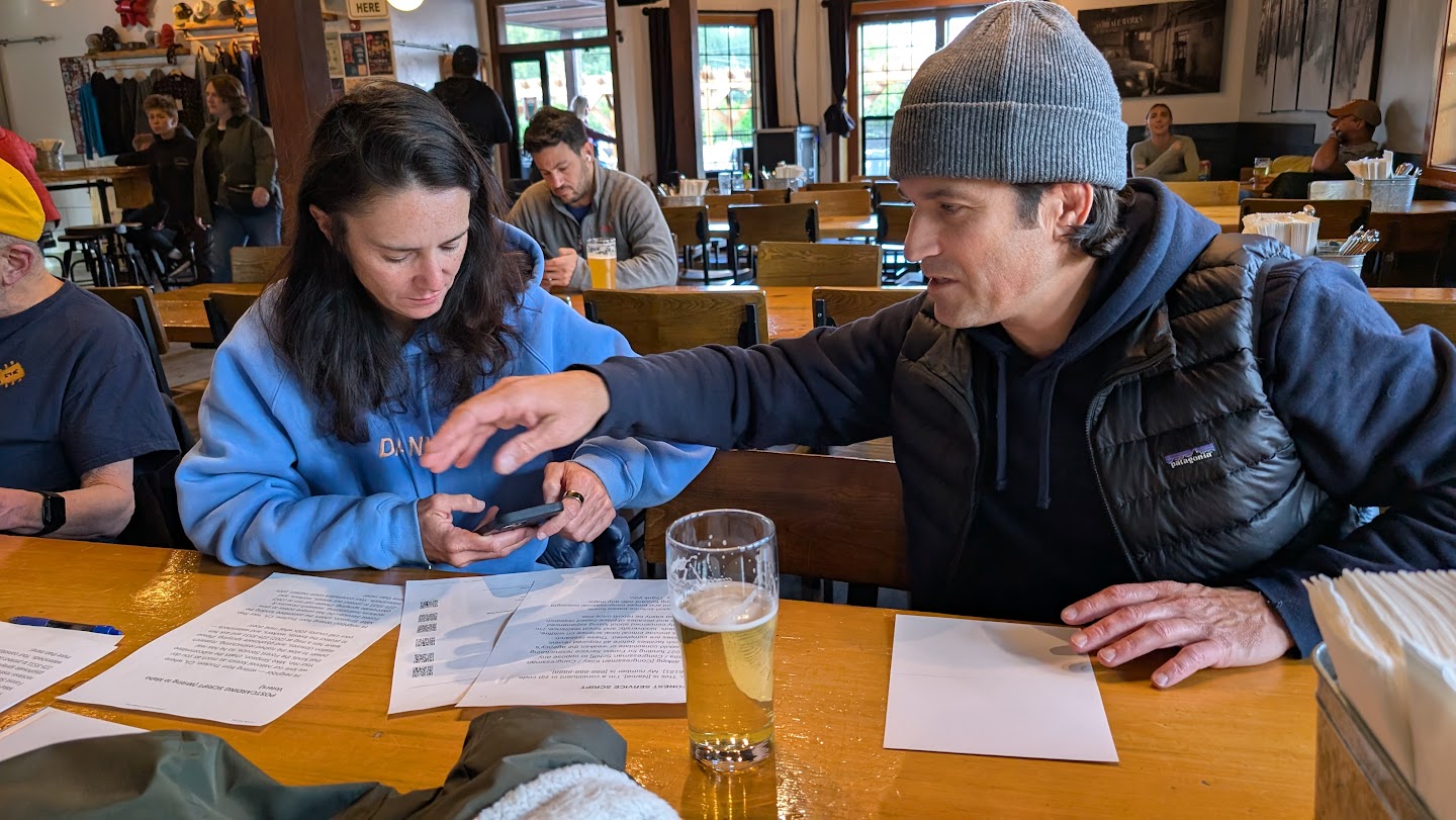 Two neighbors at a brewery table, one helping the other make a call to Congress