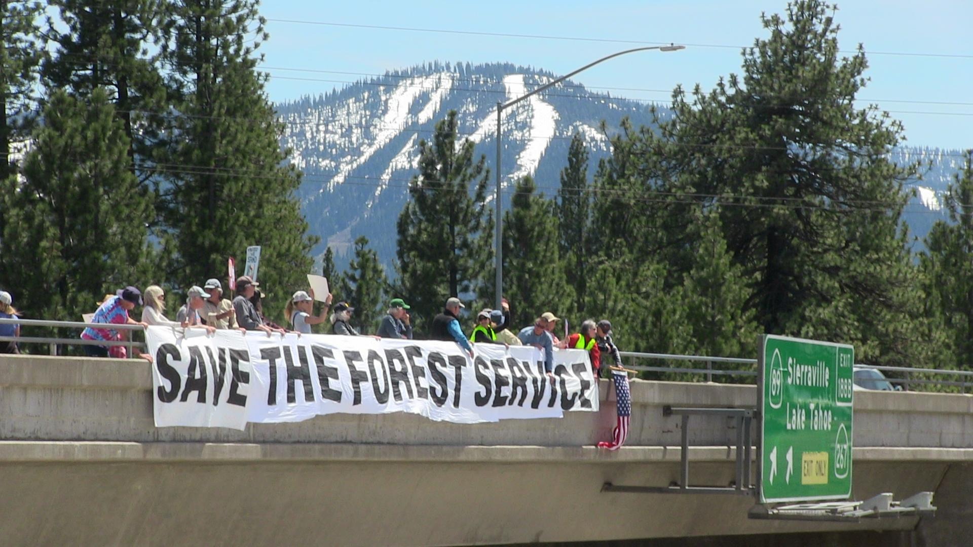 Community members hold a 'Save the Forest Service' banner on a highway overpass with the Sierra Nevada mountains in the background